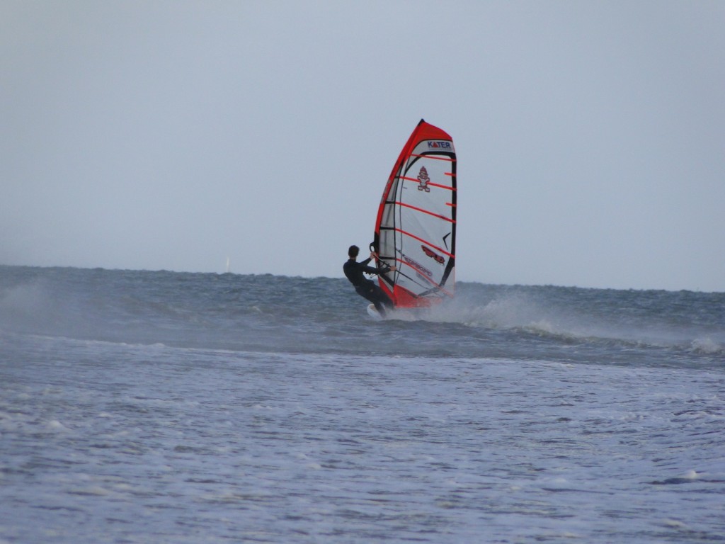 A windsurfer in a wetsuit riding a red and white sail across choppy grey water under an overcast sky.